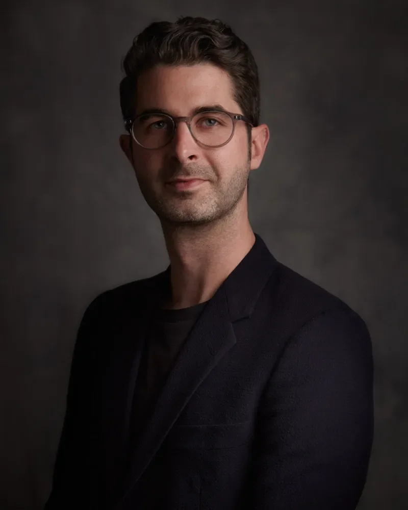 Portrait of an individual with glasses wearing a dark blazer, set against a textured grey background.