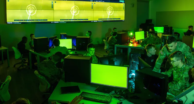 Students in military uniforms working at desktop computers in a green-lit Norwich University cybersecurity lab, with digital displays showing network diagrams.