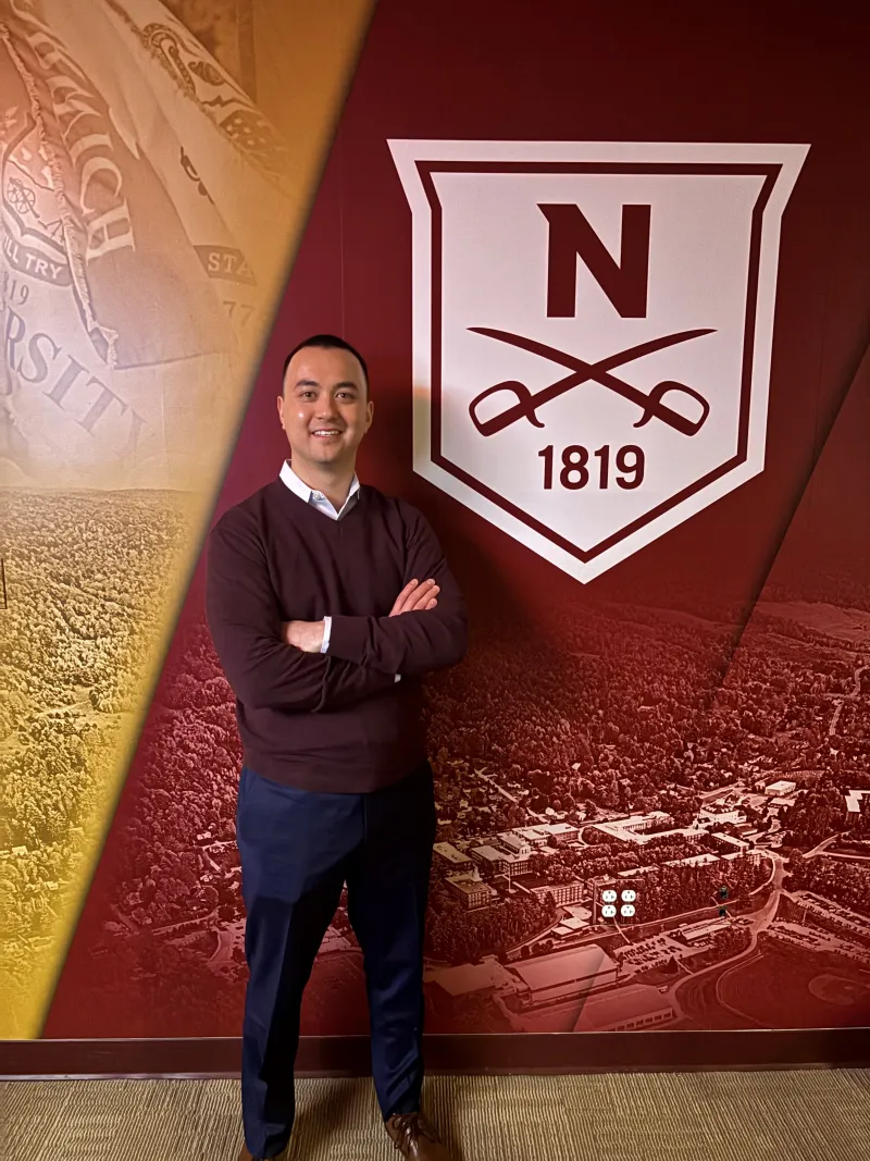 Person standing with arms crossed in front of a Norwich University banner displaying the founding year 1819 and the university emblem.