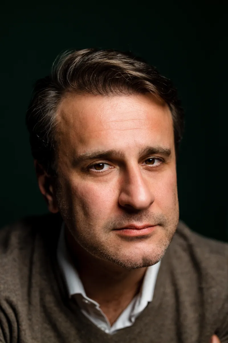 Person in a collared shirt and sweater looks toward the camera against a dark background.