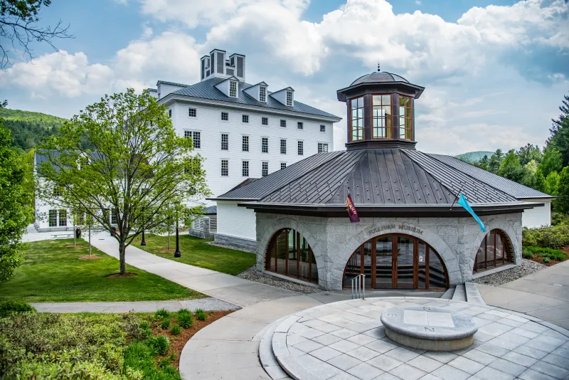 Exterior view of the Sullivan Museum and History Center at Norwich University, featuring a unique curved building with a tall cupola in front and a large multi-story building in the background, set against a backdrop of lush greenery and clear skies.