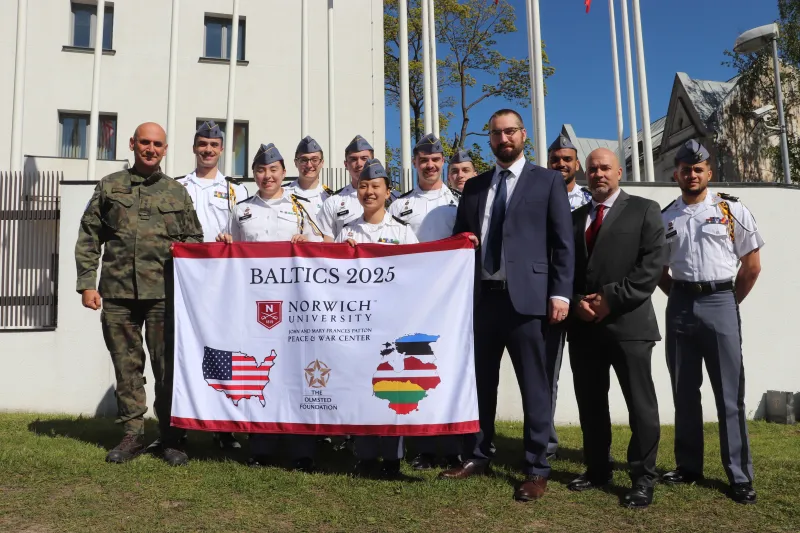 A group photo of the Olmsted trip group, holding a branded Norwich/Olmsted flag in uniform in front of military academy.