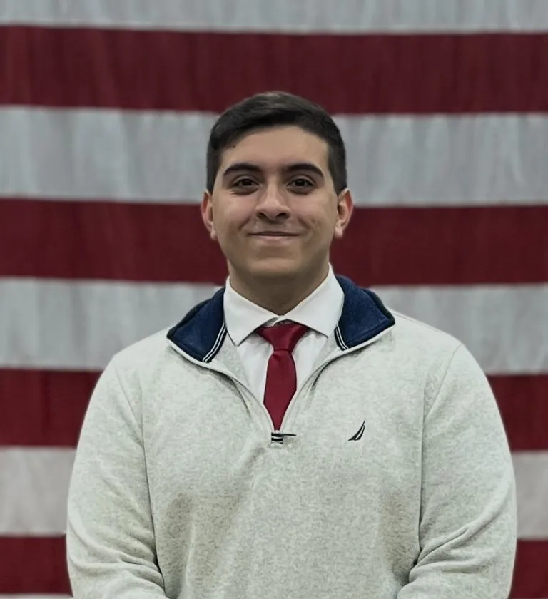 A portrait headshot of Hector Aponte in a sweater, shirt and tie in front of American flag.