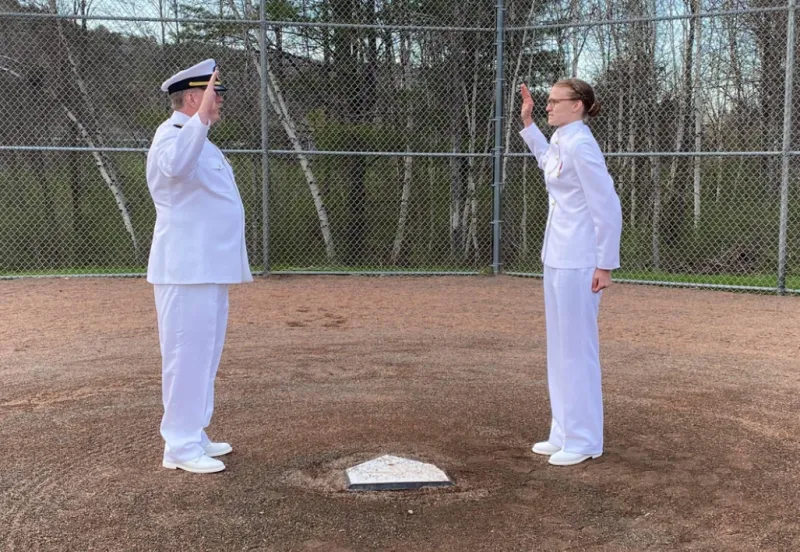 Caitlin Dailey (right) stands next to home plate while her father (left, directly across home plate) swears her in. They are both in full white Navy outfits.