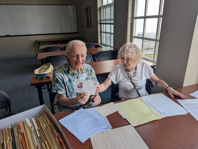 Sergey (left) in a Hawaiian styled shirt holds a letter while sitting at a table, while his wife Anna (right) points at the letter while smiling. Papers are strewn across the table.