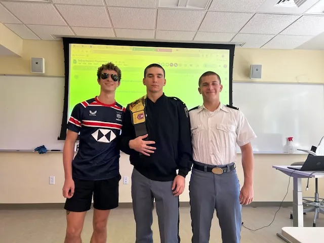 Three young men are standing together in a classroom, posing for a photo. The person on the left is wearing sunglasses and a sports jersey, the person in the middle is in a dark jacket with a patch on the shoulder, and the person on the right is wearing a white uniform shirt with gray trousers, typical of a military or cadet setting. A projection screen and whiteboards are visible in the background, suggesting this was taken during or after a class or presentation.