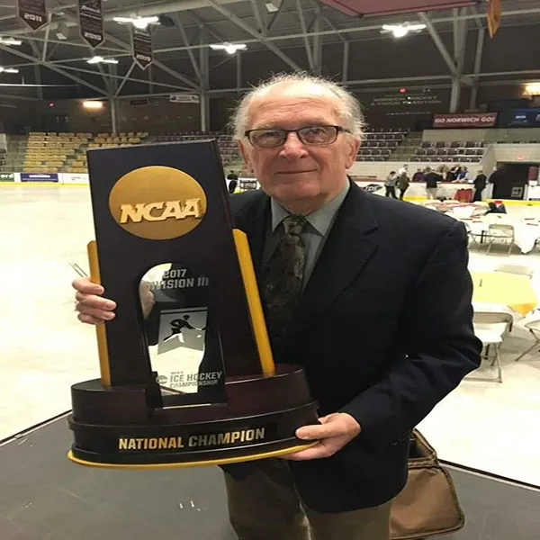 A man in a suit stands with the NCAA hockey trophy on an ice rink.