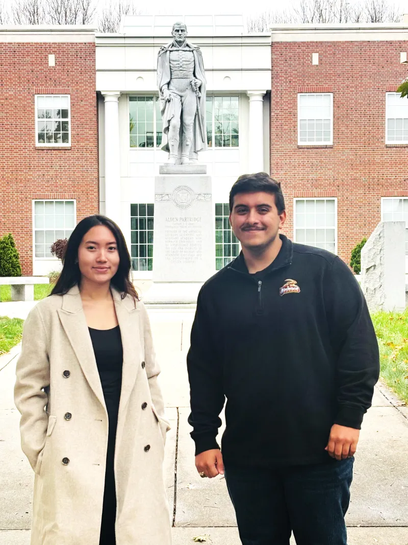 Two people stand outside near a statue in front of a red brick building with white columns.