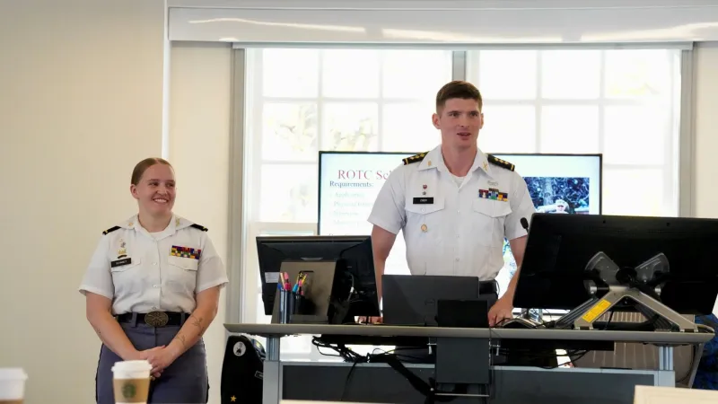 Two cadets in uniform are standing at the front of a classroom giving a presentation. One is speaking while the other smiles and listens. Both are wearing white shirts with rank insignia and ribbons. Behind them, a screen displays a slide with “ROTC Scholarship Requirements” listed. The environment appears professional and academic, with computers, notes, and coffee cups visible on the desks.