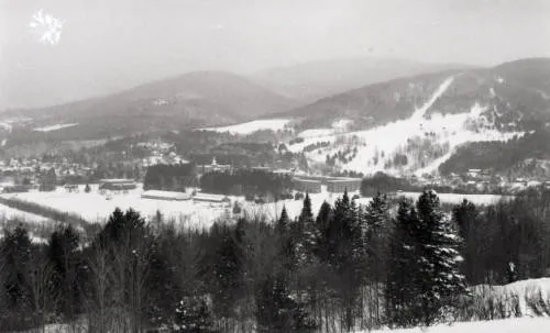 A snowy valley stretches across the scene, surrounded by forested hills and distant mountains. A small cluster of buildings sits nestled near the center, with open fields and ski trails visible along the slopes.