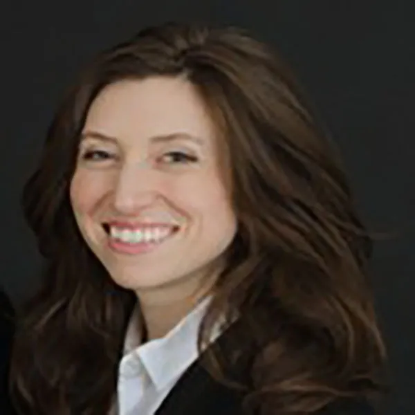 Person with long brown hair wearing a white shirt and dark jacket smiles against a dark background.