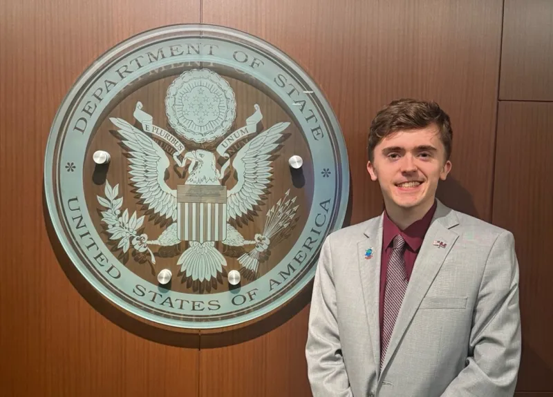 A student in a grey suit stands next to the Department of State logo.