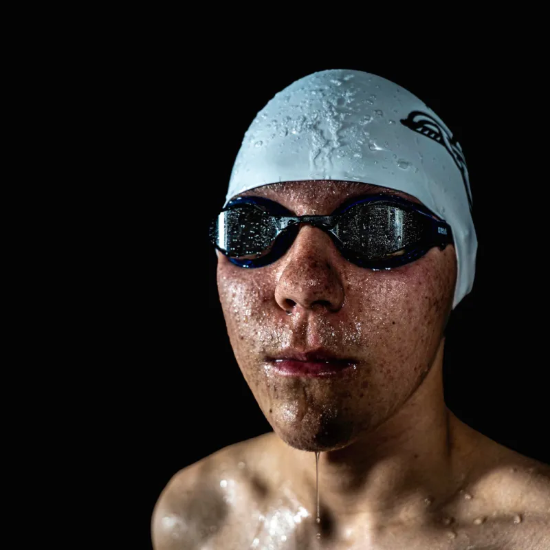 A swimmer wearing a Speedo cap and goggles, with water droplets on their face, against a dark background.