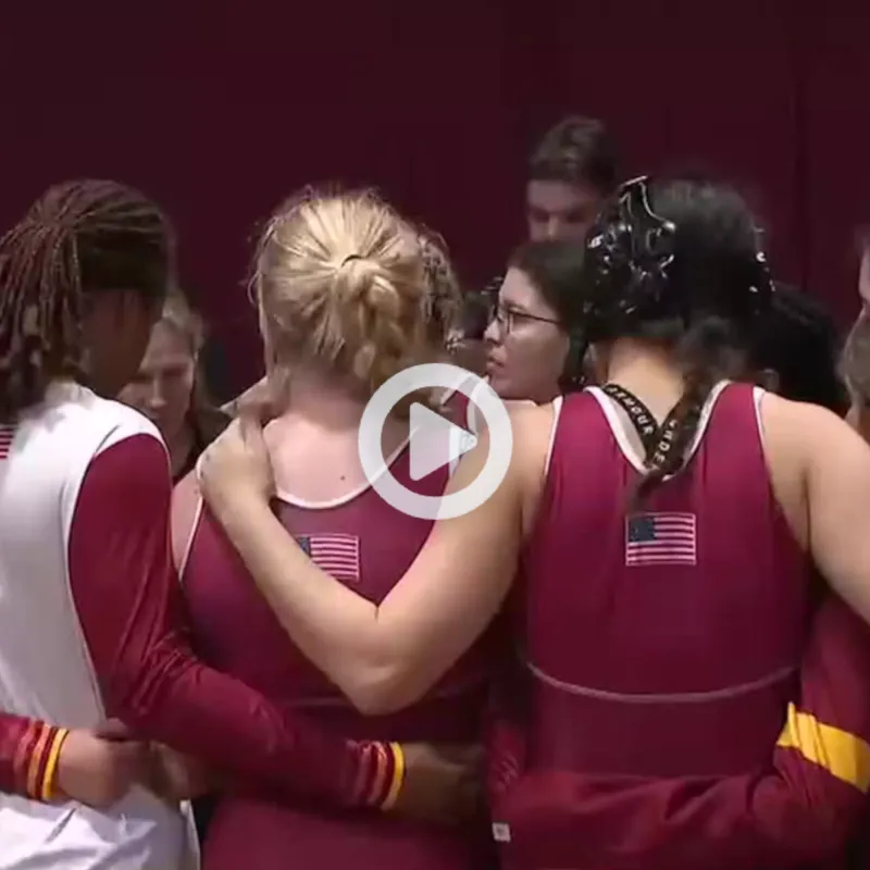 Group of athletes in maroon and gold uniforms with USA flags on their backs huddling together in a gymnasium.