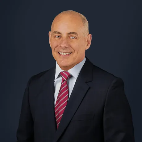 A person in a dark suit with a white shirt and red striped tie stands against a dark background.