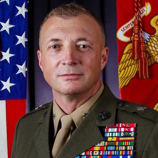 Person in a decorated military uniform stands before U.S. and Marine Corps flags.