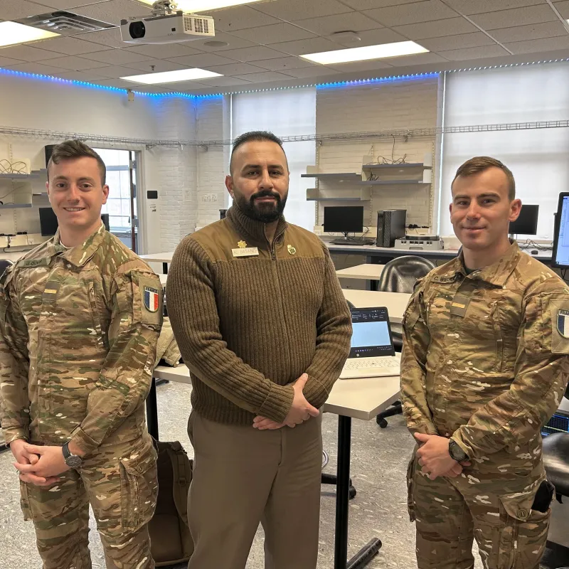 Two cadets and an instructor stand in a computer lab classroom.