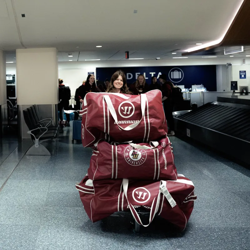 Student pushes cart stacked with Norwich University duffel bags through an airport baggage claim area.