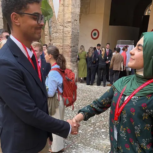Two students wearing red lanyards shake hands in a stone courtyard.