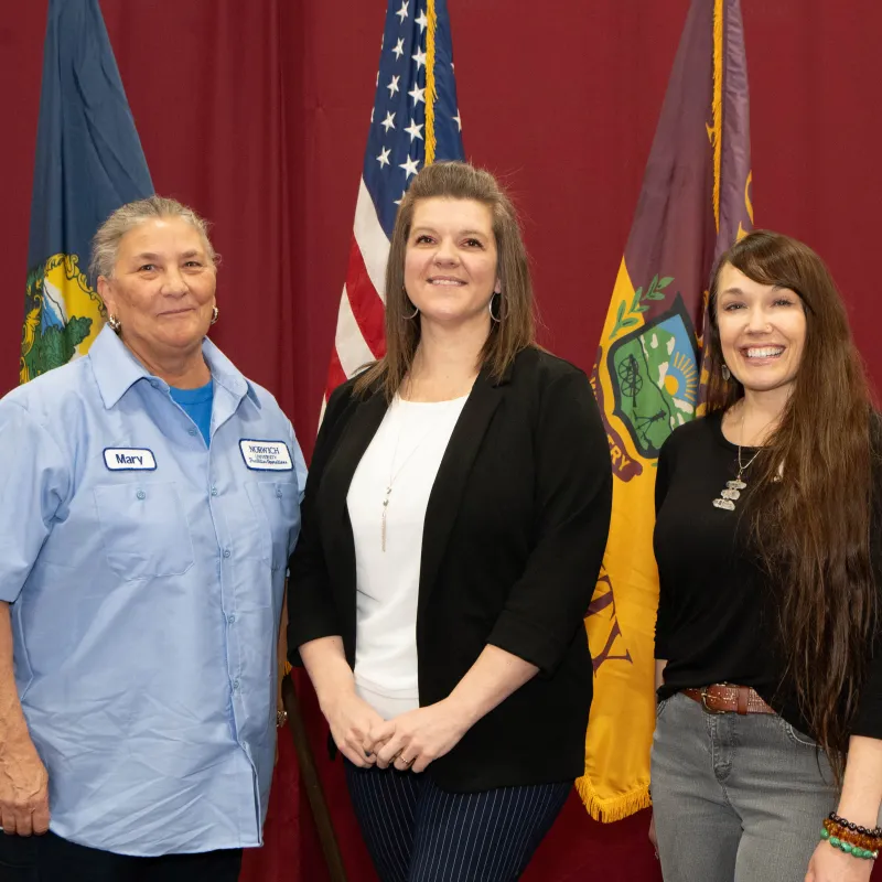 Three staff award recipients hold Norwich University certificates in front of flags.
