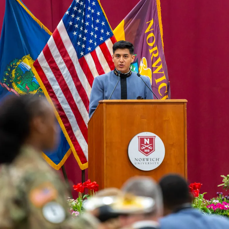 Cadet speaks at a Norwich University podium with American, Vermont, and Norwich flags behind.