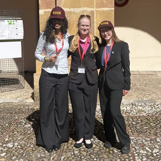 Three students wearing ROOK hats and conference badges stand in front of a stone column outdoors.