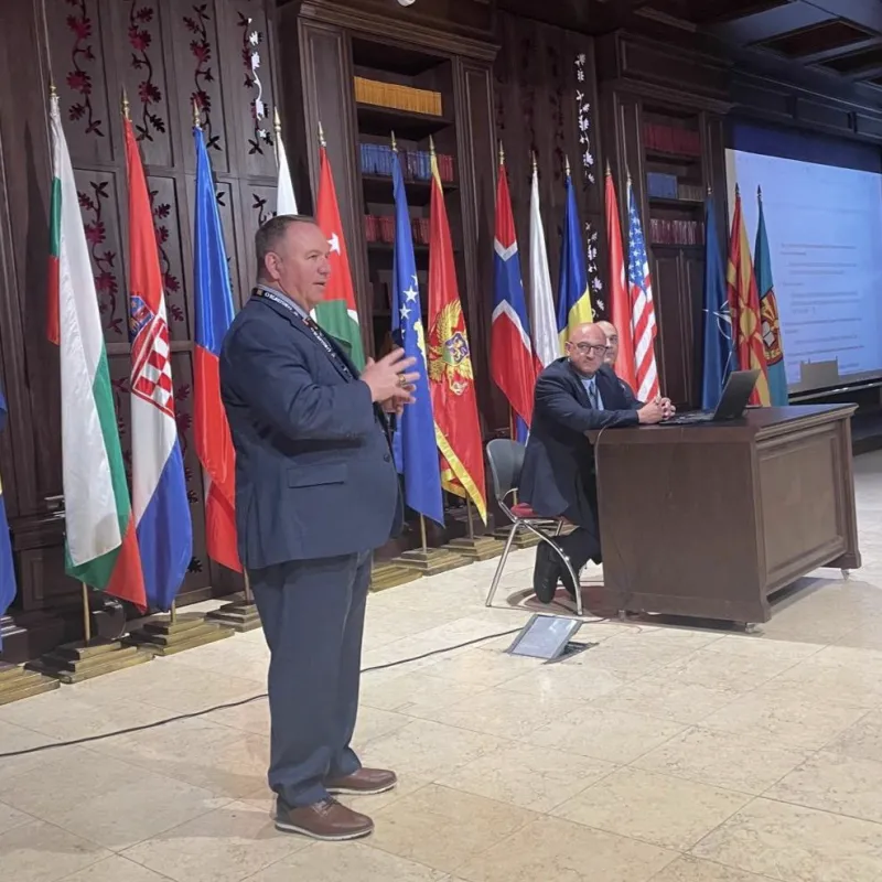 A speaker addresses an audience beside seated panelists with national flags displayed across the stage.