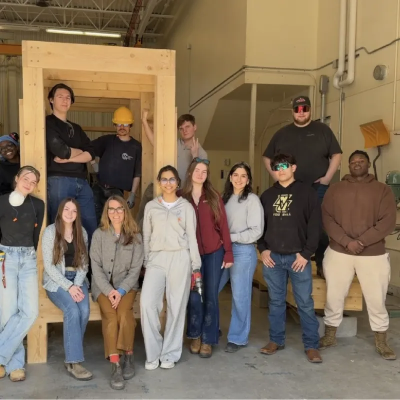 Students and an instructor pose beside a framed wood structure inside a construction lab