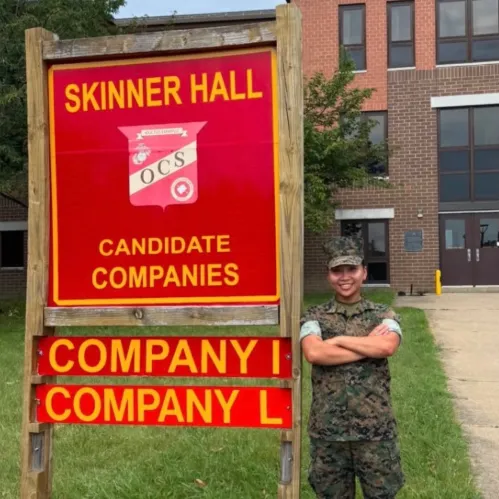 A person in camouflage uniform stands beside a Skinner Hall sign for candidate companies I and L.