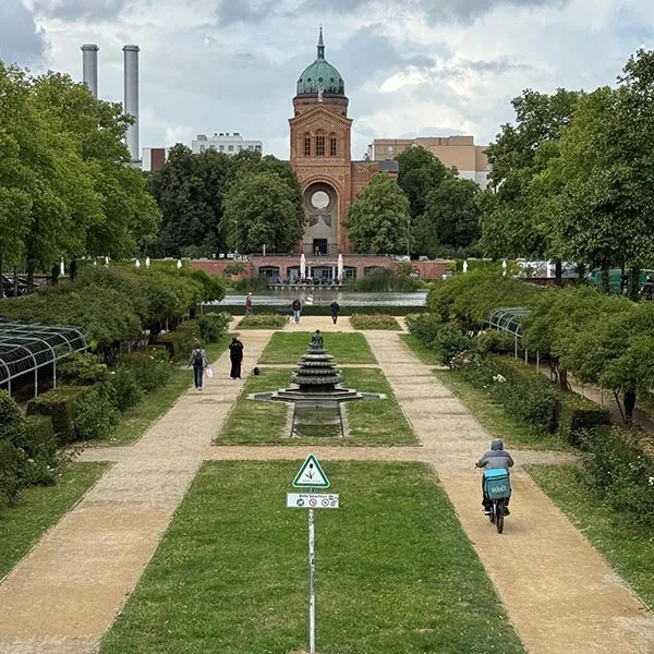 Formal garden with a central fountain, walking paths, trees, cyclists, and a brick tower in the distance.