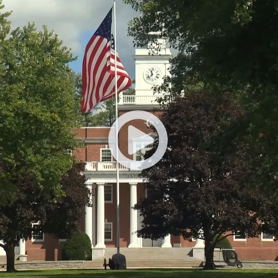American flag flies in front of a brick building with white columns and a clock tower.