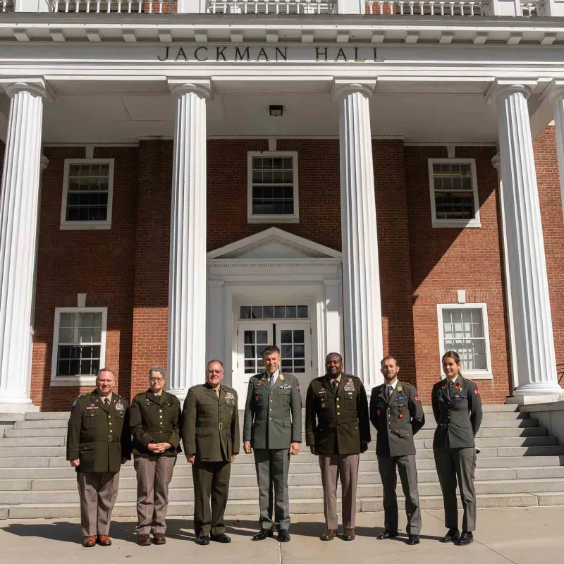 Seven service members in military dress uniforms stand on the steps in front of Jackman Hall at Norwich University.